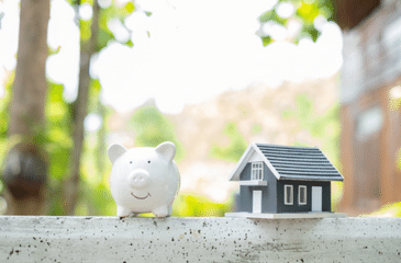 A white piggy bank and a small model home are placed side by side on a ledge, symbolizing the concept of savings and investment in real estate.